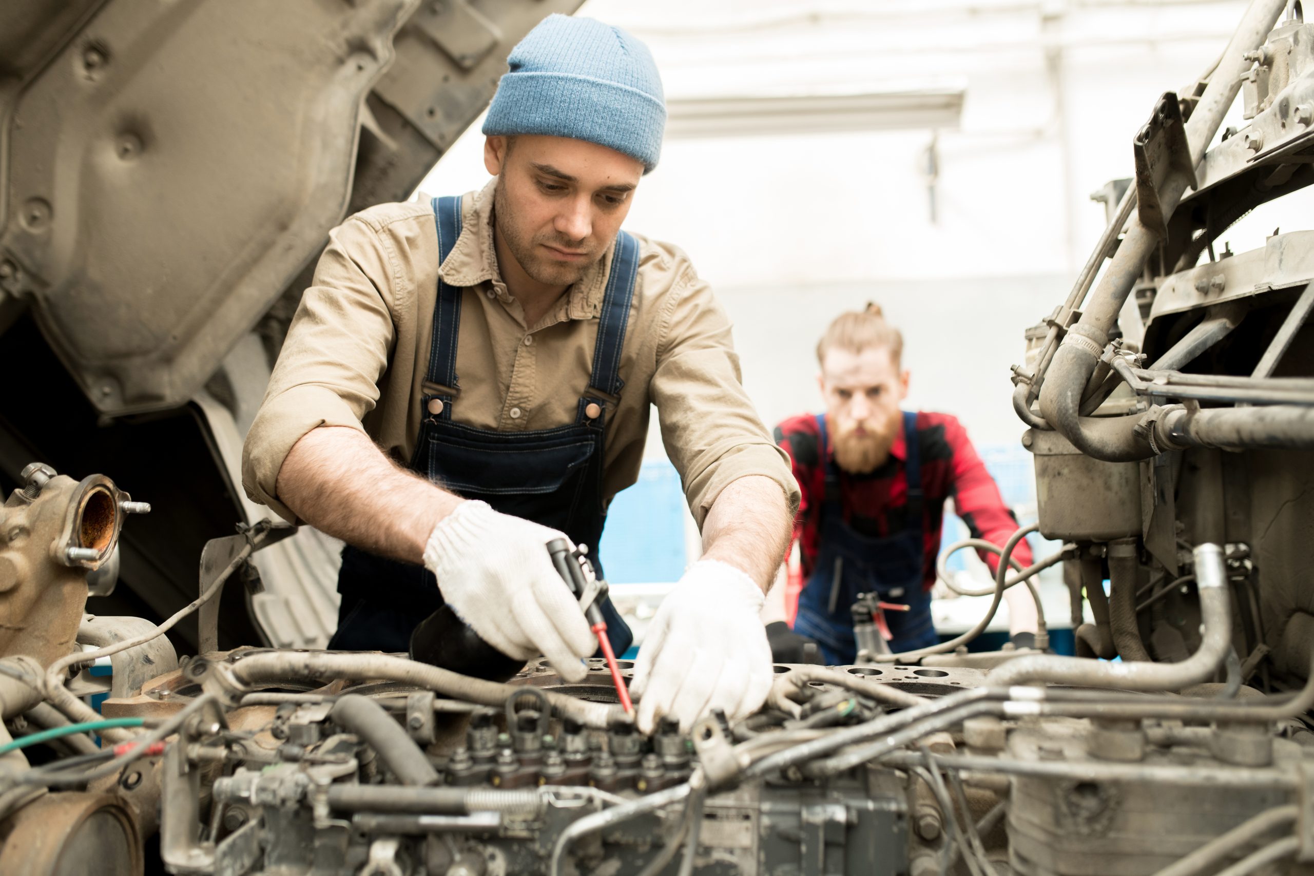 Young concentrated Caucasian male technician repairing vehicle in service garage, male colleague watching him in background